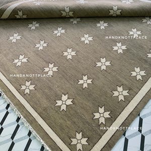 May include: A rolled-up, rectangular rug with a repeating starburst pattern. The rug is primarily a muted olive green with a cream-colored border and starbursts. The text "HANDKNOTTPLACE" is visible on the rug.
