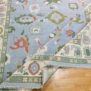 K&ouml;nnte beinhalten: Hellblauer Teppich mit floralem Muster in Gr&uuml;n-, Rot- und Graut&ouml;nen. Der Teppich hat einen cremefarbenen Rand mit einem sich wiederholenden Blumendesign und dem Text "HANDKNOTPLACE". Der Teppich ist teilweise aufgerollt und zeigt die Unterseite.