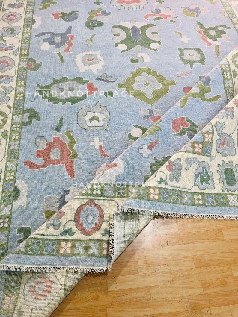 K&ouml;nnte beinhalten: Hellblauer Teppich mit floralem Muster in Gr&uuml;n-, Rot- und Graut&ouml;nen. Der Teppich hat einen cremefarbenen Rand mit einem sich wiederholenden Blumendesign und dem Text "HANDKNOTPLACE". Der Teppich ist teilweise aufgerollt und zeigt die Unterseite.