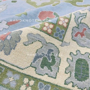 K&ouml;nnte beinhalten: Nahaufnahme eines handgekn&uuml;pften Teppichs mit hellblauem Hintergrund und detaillierten Blumenmustern in Gr&uuml;n, Rosa und Grau. Das Design des Teppichs weist einen Rand mit sich wiederholenden Blumenmotiven und ein cremefarbenes Mittelfeld auf.