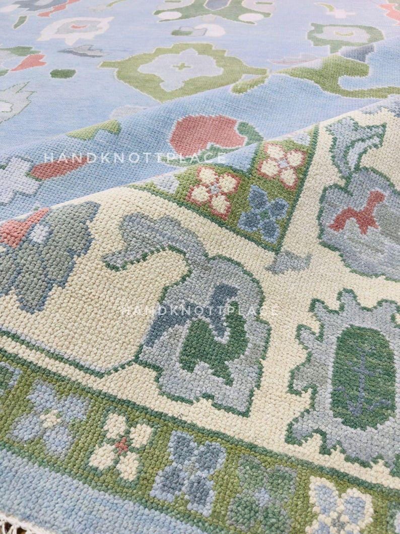K&ouml;nnte beinhalten: Nahaufnahme eines handgekn&uuml;pften Teppichs mit hellblauem Hintergrund und detaillierten Blumenmustern in Gr&uuml;n, Rosa und Grau. Das Design des Teppichs weist einen Rand mit sich wiederholenden Blumenmotiven und ein cremefarbenes Mittelfeld auf.