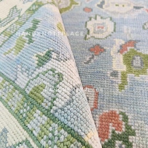 Op de afbeelding: Close-up van een opgerold vloerkleed met een lichtblauwe achtergrond en een bloemenpatroon in groen, wit en koraal. Het kleed heeft een getextureerd oppervlak en de tekst "HANDKNOTTPLACE" is zichtbaar.
