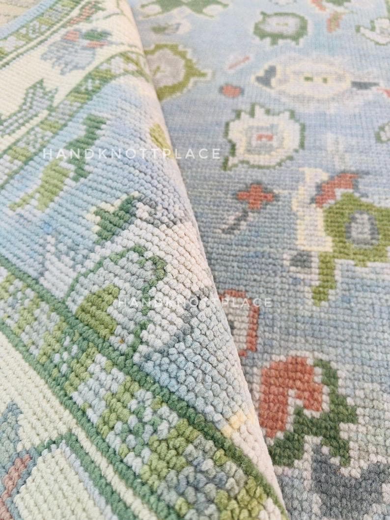 K&ouml;nnte beinhalten: Nahaufnahme eines aufgerollten Teppichs mit hellblauem Hintergrund und einem Blumenmuster in Gr&uuml;n-, Wei&szlig;- und Korallt&ouml;nen. Der Teppich hat eine strukturierte Oberfl&auml;che und der Text "HANDKNOTTPLACE" ist sichtbar.