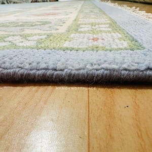 K&ouml;nnte beinhalten: Nahaufnahme eines rechteckigen Teppichs mit hellblauem Rand und einem zentralen Design mit gr&uuml;nen und orangefarbenen Blumenmustern. Der Teppich hat einen dicken, strukturierten Flor und liegt auf einem Holzboden.