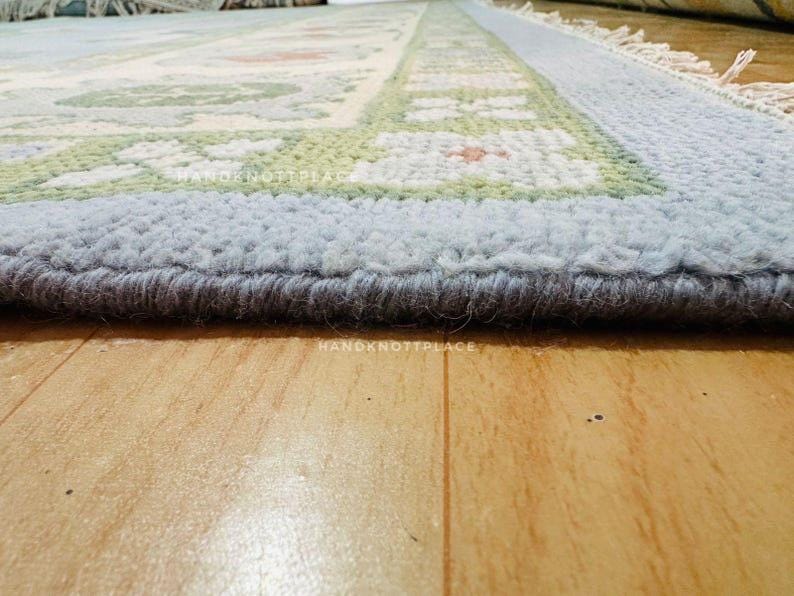 K&ouml;nnte beinhalten: Nahaufnahme eines rechteckigen Teppichs mit hellblauem Rand und einem zentralen Design mit gr&uuml;nen und orangefarbenen Blumenmustern. Der Teppich hat einen dicken, strukturierten Flor und liegt auf einem Holzboden.