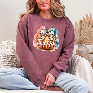 May include: A maroon sweatshirt featuring a colorful pumpkin graphic with a polka dot bow and the word "Thankful" in white script. The pumpkin is set against a backdrop of orange, teal, and pink brushstrokes, creating a festive autumn design.