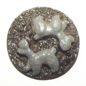 May include: A circular button featuring a dark brown, textured background with a pebbled surface. Two raised, light gray poodle figures are positioned on the surface, one standing near the bottom and one positioned diagonally above it.