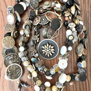 Puede incluir: Una colección de botones antiguos de varios tamaños, formas y colores, incluyendo negro, dorado, plateado y nacarado. Algunos botones presentan diseños y adornos intrincados. Los botones están dispuestos sobre una superficie de madera.