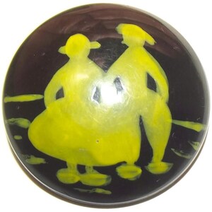 May include: A circular, dark brown button featuring a bright yellow, stylized graphic of two figures standing side-by-side. The figures are depicted in a simple, abstract form with rounded shapes, set against the smooth, dark background of the button.