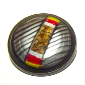 May include: A circular bronze button with a striped interior. The button has a central rectangular design with a mosaic of gold, white, red, and yellow colours. The button is set against a white background.