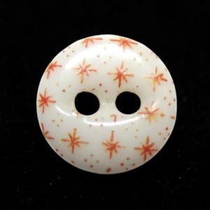 May include: A white button with two holes and a pattern of orange starbursts and small dots. The button is round and has a glossy finish, suitable for sewing or craft projects.