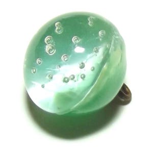 May include: A translucent, oval-shaped green glass button with embedded air bubbles. The button has a brass-colored shank on the back for attachment. The glass has a slight iridescence, and the bubbles vary in size.
