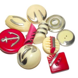 May include: A collection of assorted buttons in various shapes and sizes, featuring cream, red, and pink colors with metallic silver accents. The set includes round, square, and elongated designs, some with anchor motifs, ridges, and smooth surfaces.