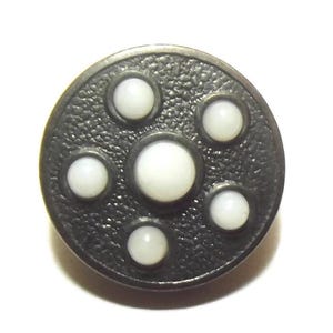 May include: A round, dark gray button with a textured surface. The button features six white, circular accents arranged in a pattern. The central accent is larger than the surrounding five.