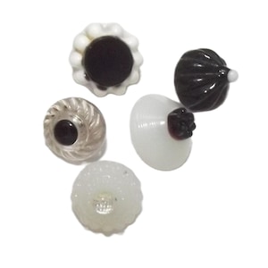 Può includere: Una collezione di cinque bottoni decorativi in vetro di varie forme e colori. I bottoni includono un bottone bianco e nero a forma di fiore, un bottone trasparente con un centro nero e un bottone bianco con un centro rosso scuro.