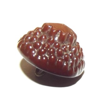 Puede incluir: Un botón vintage marrón oscuro con una superficie texturizada. El botón tiene una forma redondeada con una serie de pequeñas protuberancias elevadas que cubren la superficie. Un lazo de metal es visible en la parte inferior.