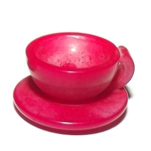 May include: A solid, bright pink miniature cup and saucer set. The cup features a rounded bowl shape with a small, curved handle attached to the side, resting securely on a matching circular saucer. The entire piece has a smooth, uniform finish.