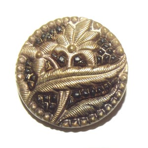 May include: A detailed, antique button with a gold-toned finish. The button features an intricate design of leaves and geometric shapes, with small black accents. The edge is lined with tiny, raised dots. A vintage sewing notion.