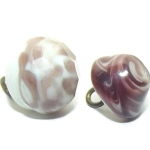 May include: Two vintage earrings. One is a round shape with white and brown swirls, the other is a heart shape with dark purple swirls. Both have a gold-colored loop.
