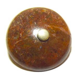 Può includere: Una pietra rotonda e lucidata con un motivo screziato di tonalità marroni, rosse e beige. Un piccolo borchia metallica color argento è incastonata sulla superficie superiore. La pietra ha una finitura liscia e lucida, suggerendo un oggetto decorativo.