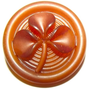 May include: A round, amber-colored button featuring a raised, three-leaf clover design in the center. The button has a smooth, glossy finish with concentric circular grooves surrounding the clover motif, creating a textured appearance.