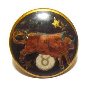 Puede incluir: Un botón redondo de estilo vintage con un borde dorado. El botón presenta un toro marrón con cuernos azules sobre un fondo negro, con detalles de estrellas doradas y el símbolo de Tauro. El botón probablemente es de metal o esmalte.