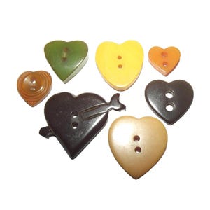 May include: A collection of heart-shaped buttons in various colors, including green, yellow, orange, brown, and black. Some buttons have two holes, while others have a single hole. One button features an arrow design.