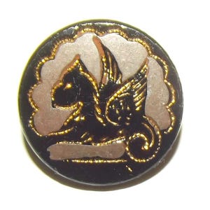 May include: A round black button featuring a central embossed design of a winged creature in profile. The figure is highlighted with gold-colored metallic paint and set against a light gray, cloud-like background within the circular frame.