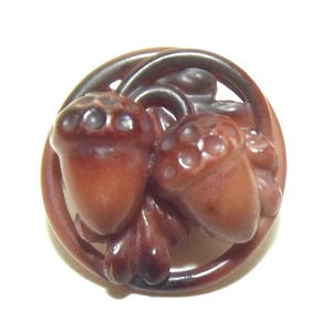 Puede incluir: Botón tallado marrón detallado con dos bellotas y hojas dentro de un marco circular. Las bellotas tienen tapas texturizadas y son de color marrón rojizo. El botón probablemente está hecho de un material duro.