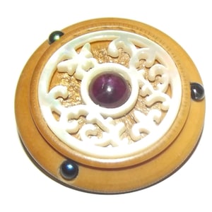 May include: A circular brooch featuring a tan base with three small dark blue spherical accents along the edge. The center contains a white carved circular inlay with an intricate openwork pattern surrounding a single smooth dark purple cabochon.