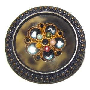 May include: A round, ornate button with a dark gray border and a central design. The center features a gold-colored floral pattern with various colored gems, including blue, green, and red. The button's outer edge has small, evenly spaced gold-colored studs.