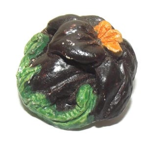 Op de afbeelding: Een keramische sculptuur van een donkerbruine, afgeronde vorm met groene, bladachtige accenten en een oranje bloem. De sculptuur heeft een glanzende afwerking en staat tegen een witte achtergrond.