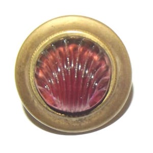 Puede incluir: Un botón redondo y antiguo con un marco de metal color latón. El centro presenta un diseño translúcido en forma de concha en tonos rojos y rosas. El botón está sobre un fondo blanco.