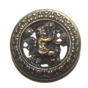 Può includere: Bottone antico in bronzo con un rilievo dettagliato di un cherubino che suona uno strumento e due sfere. Il cherubino è all'interno di una cornice circolare con un motivo floreale ornamentale ripetuto. Il bottone ha un aspetto invecchiato.