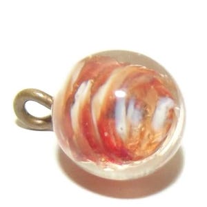 Puede incluir: Una cuenta de vidrio redonda y transparente con un remolino de colores rojo, blanco y dorado en su interior. Tiene un pequeño lazo de color bronce adjunto en la parte superior. La cuenta está sobre un fondo blanco.