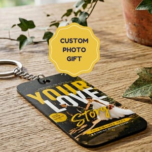 Op de afbeelding: Een rechthoekige sleutelhanger met een aangepaste foto van een persoon in een witte jurk, met de vetgedrukte gele en witte tekst "YOUR LOVE STORY" op een donkere achtergrond, bevestigd aan een zilveren metalen ring en ketting.