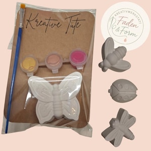 Könnte beinhalten: Ein Bastelset mit einem weißen Schmetterling, drei Farbtöpfen und einem Pinsel. Das Set ist in einer transparenten Plastiktüte verpackt, mit dem Text "Kreative Tüte" auf einer braunen Karte. Zusätzliche unbemalte Insektenfiguren sind enthalten.