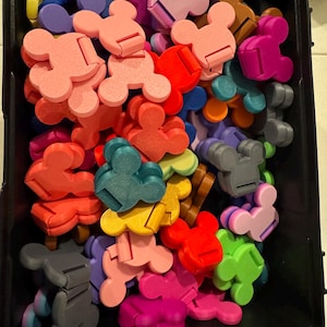 Può includere: Una collezione di piccoli supporti per telefono in plastica multicolore a forma di teste di topo. I supporti sono sparsi all'interno di un contenitore rettangolare nero e presentano vari colori vivaci tra cui rosa, rosso, blu, giallo, verde e grigio.