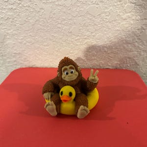 May include: A brown, furry, ape-like figurine with a smiling face, sitting inside a bright yellow rubber duck floatie. The figurine has one hand raised in a peace sign gesture, with visible toes and fingers, and the duck has a distinct orange beak.