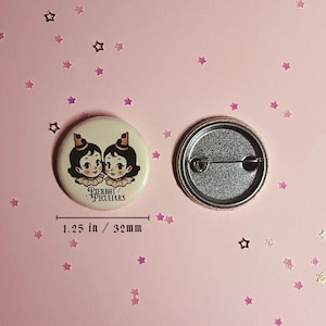 May include: A round pinback button measuring 1.25 inches in diameter. The front features an illustration of two identical characters with dark hair, small hats, and ruffled collars, with the text Pierrot Peculiars. The back shows a metal pin mechanism.