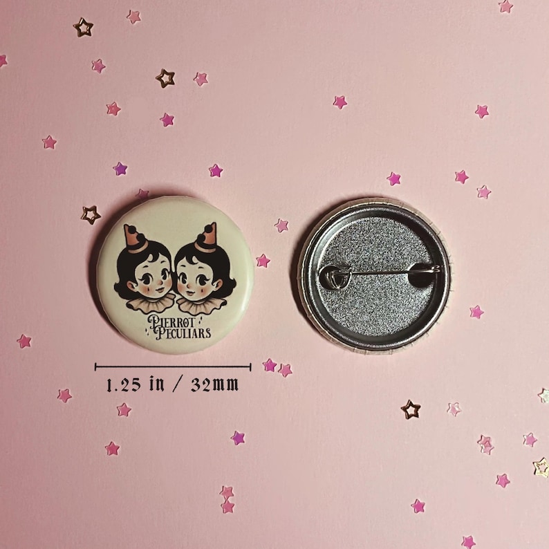 May include: A round pinback button measuring 1.25 inches in diameter. The front features an illustration of two identical characters with dark hair, small hats, and ruffled collars, with the text Pierrot Peculiars. The back shows a metal pin mechanism.
