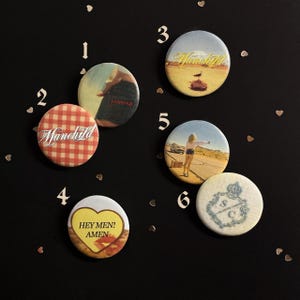 May include: Six circular pinback buttons featuring various designs: a red and white gingham pattern with the text Manchild, a denim pocket, a desert landscape with a bird, a person hitchhiking, a heart with the text HEY MEN! AMEN, and a blue crest.