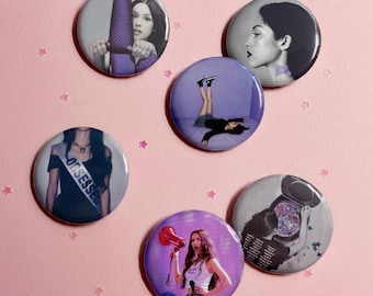 May include: A set of five circular pin-back buttons featuring various photographic images of a person. One button shows a person wearing a sash that reads OBSESSED. Other buttons depict a person with a megaphone, lying on a floor, or posing.