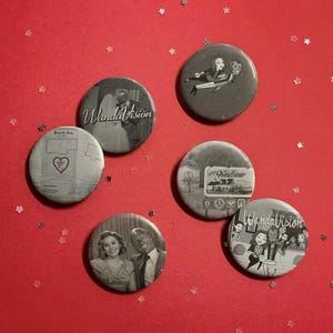 May include: A collection of six round, grayscale buttons with images and text from the show "WandaVision." The buttons feature scenes and the show's logo, set against a red background with star-shaped confetti.