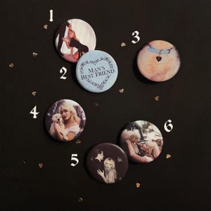 May include: A collection of six circular pin-back buttons featuring various photographic prints. One button is light blue with the text "MAN'S BEST FRIEND" inside a decorative heart frame. Other buttons display images of a person with a dog.