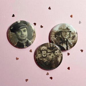 May include: Three round pinback buttons with sepia-toned images of men in military uniforms. The buttons are arranged on a light pink surface scattered with small heart-shaped confetti.
