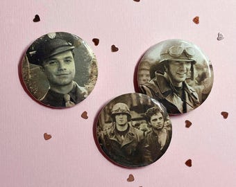 Puede incluir: Tres chapas redondas con alfiler que presentan impresiones fotográficas en tono sepia. Una chapa muestra a un hombre con gorra militar, otra a un soldado con gafas protectoras y la tercera representa a dos hombres, uno con casco.