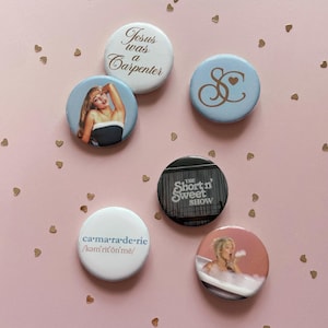 May include: Six circular pinback buttons featuring various designs: one white with the text 'Jesus was a Carpenter', one light blue with a gold 'SC' monogram, one with a portrait of a person, one black with 'THE SHORT N' SWEET SHOW' text, one white with 'ca·ma·ra·de·rie' text, and one with a person in a bathtub.