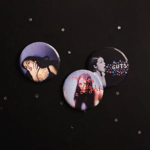 May include: Three circular pin-back buttons featuring photographic imagery. One button shows a person with dark hair, another displays the text GUTS with colorful stars, and the third features the text bad idea right with a portrait.