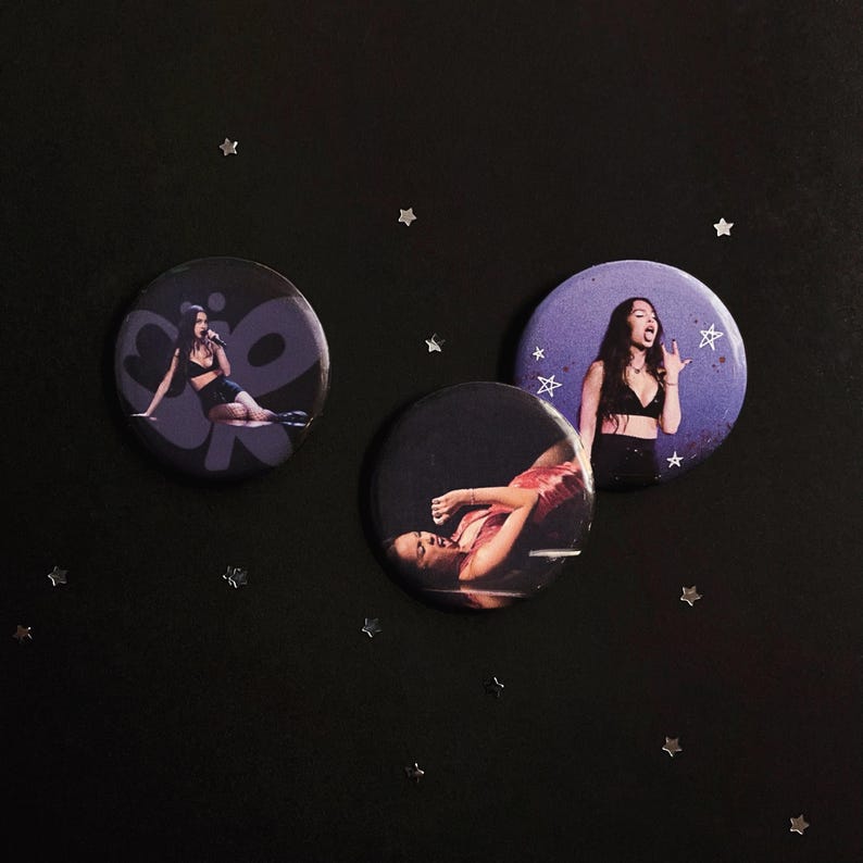 May include: Three circular pin-back buttons featuring photographic prints of a person with long dark hair. One button shows a person in a black top, another shows a person in a red top, and the third shows a person in a black top with white star graphics.